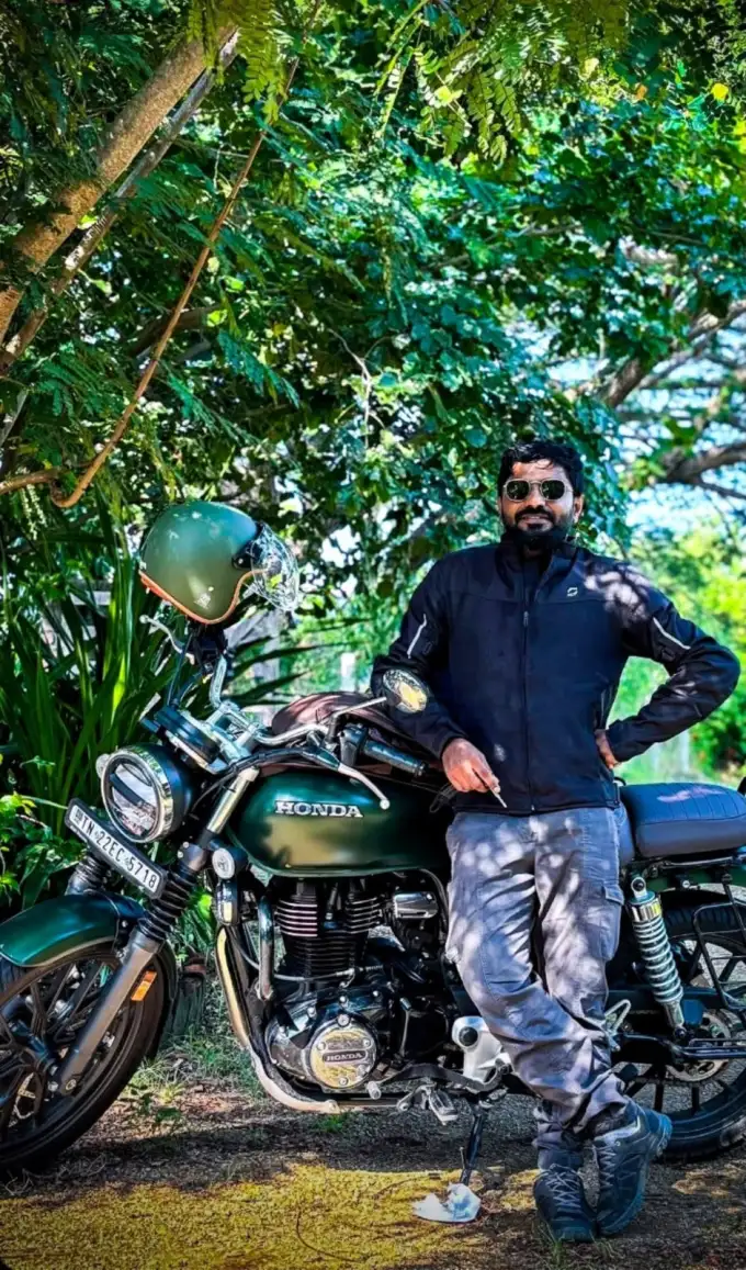 Niyas leaning on a green Honda motorcycle under trees